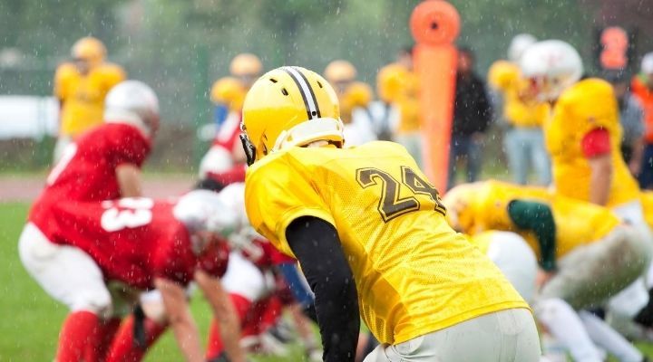Back view of a football player in yellow uniform, his hands on his knees and witnessing the game while it's raining on the field